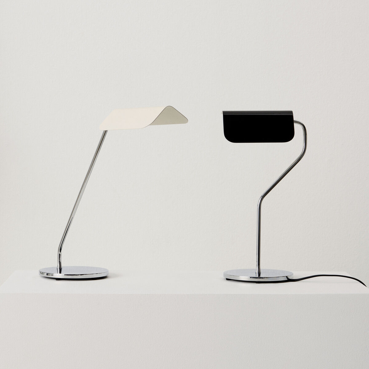 Zwei moderne Schreibtischlampen der Marke Apex Lampe »Desk« stehen auf einer weißen Oberfläche. Eine Lampe ist weiß mit einer geschwungenen Form, die andere ist schwarz mit einem minimalistischen Design. Beide haben eine stabile Metallbasis und passen zu einer zeitgemäßen Einrichtung.