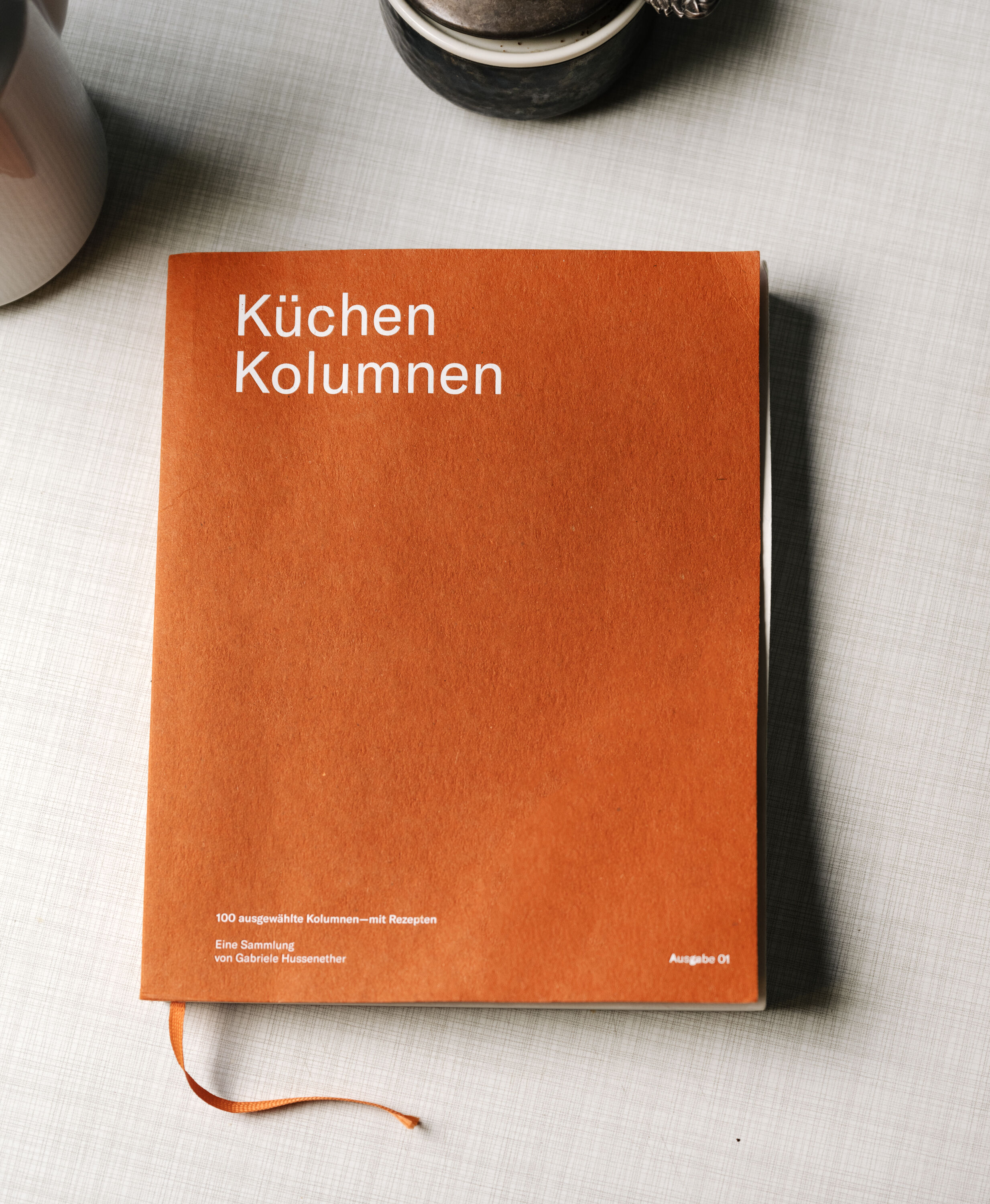 Ein orangefarbenes Buch mit dem Titel 'Küchen Kolumnen' liegt auf einer hellen Tischdecke. Das Cover zeigt weißen Text, und ein orangefarbenes Leseband ist sichtbar. Im Hintergrund sind unscharf eine Tasse und der Rand eines Gegenstands zu erkennen.