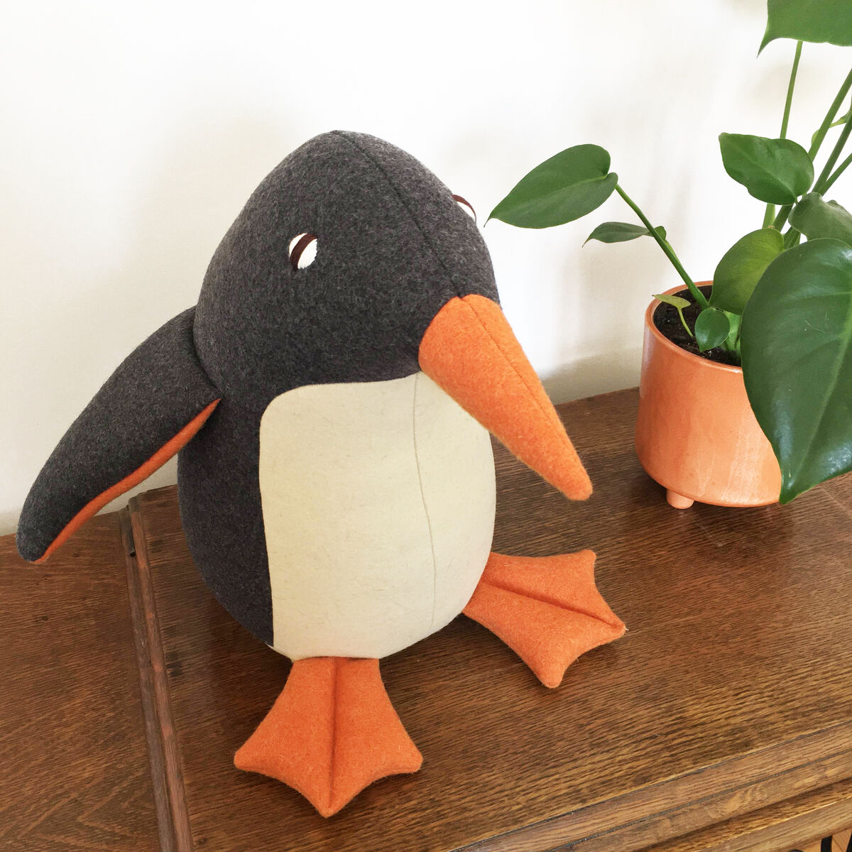 Ein handgemachter Pinguin namens »Oliver« aus Wolle sitzt auf einem Holztisch neben einer Pflanze in einem orangefarbenen Blumentopf. Der Pinguin hat einen grauen Körper, orangefarbene Flügel und Füße sowie einen weißen Bauch. Die Augen sind dunkel und aus Knöpfen gefertigt.