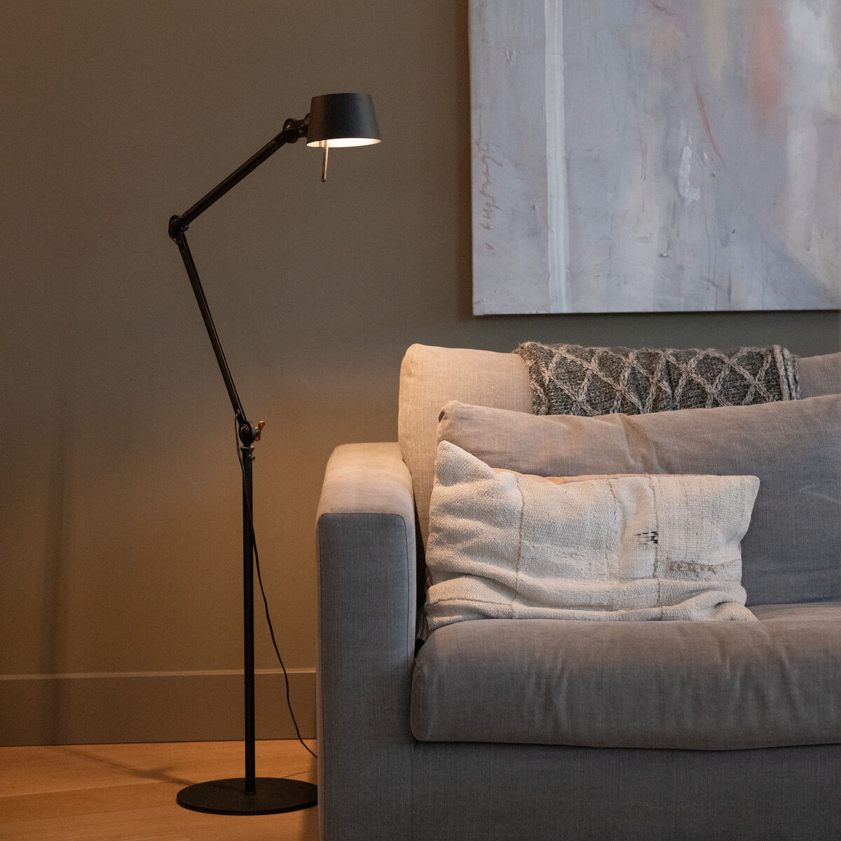 Eine moderne Stehlampe mit zwei Armen in schwarzer Farbe steht neben einer grauen Couch in einem stilvollen Wohnzimmer. Im Hintergrund ist ein abstraktes Kunstwerk an der Wand zu sehen. Die Beleuchtung schafft ein gemütliches Ambiente.