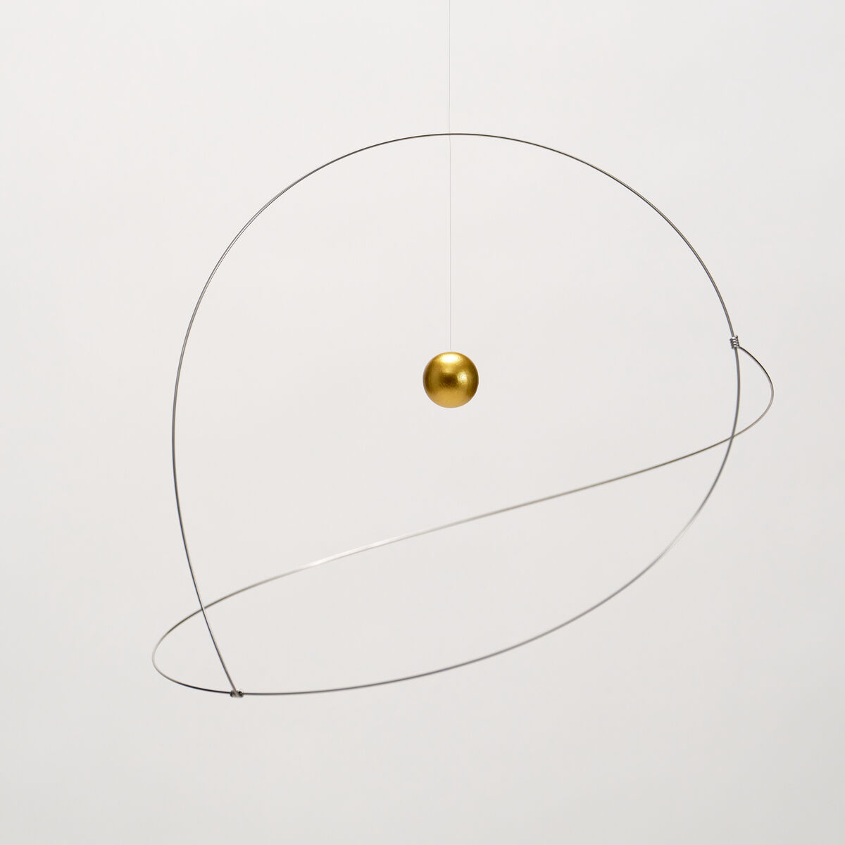 Das Mobile "Metamorphosis, Golden Planet" von Flensted Hanging Art zeigt eine goldene Kugel, die in der Mitte eines zarten silbernen Metallrings schwebt. Das Mobile hängt an einem dünnen, fast unsichtbaren Faden vor weißem Hintergrund, minimalistisch und elegant arrangiert.