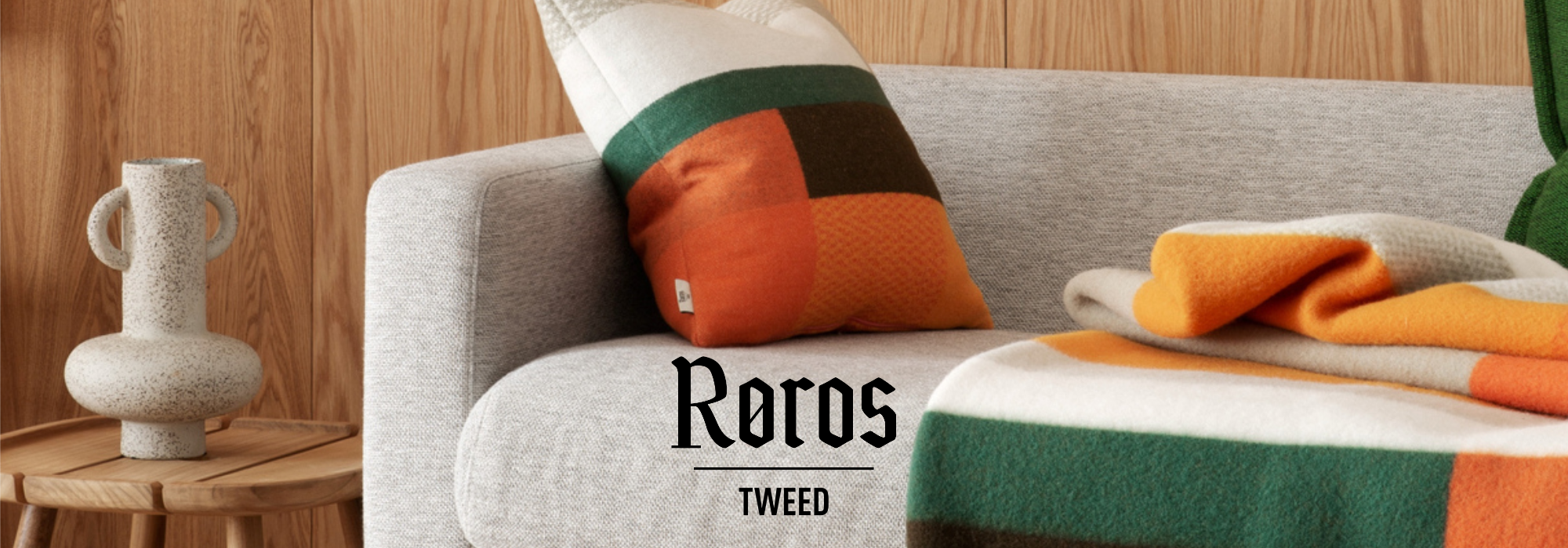 Hellgraues Sofa mit Kissen und Decke in Orange, Grün und Weiß vor Holzwand, daneben ein grauer Keramikkrug auf einem Holzbeistelltisch