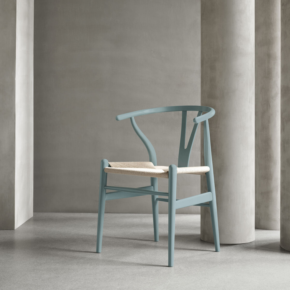 Ein moderner Stuhl, Modell Stuhl CH24 Soft von Carl Hansen, steht vor einer eleganten grauen Wand mit Bogendurchgänge. Der Stuhl hat eine blaue Lackierung mit einer naturfarbenen Sitzfläche. Minimalistisches skandinavisches Design.