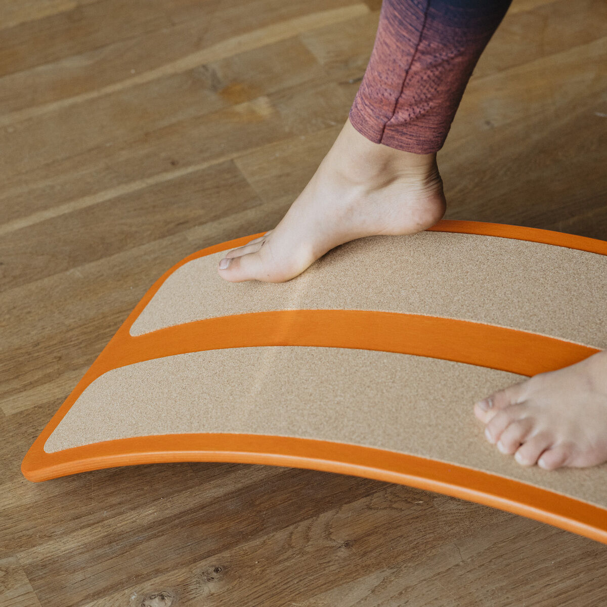 Nahaufnahme von nackten Füßen auf einem Balance Board »das.Brett« in pompejanisch rot, stehend auf einem Parkettboden. Das Balance Board zeigt eine leichte Wölbung und hat beige und rote Bereiche.