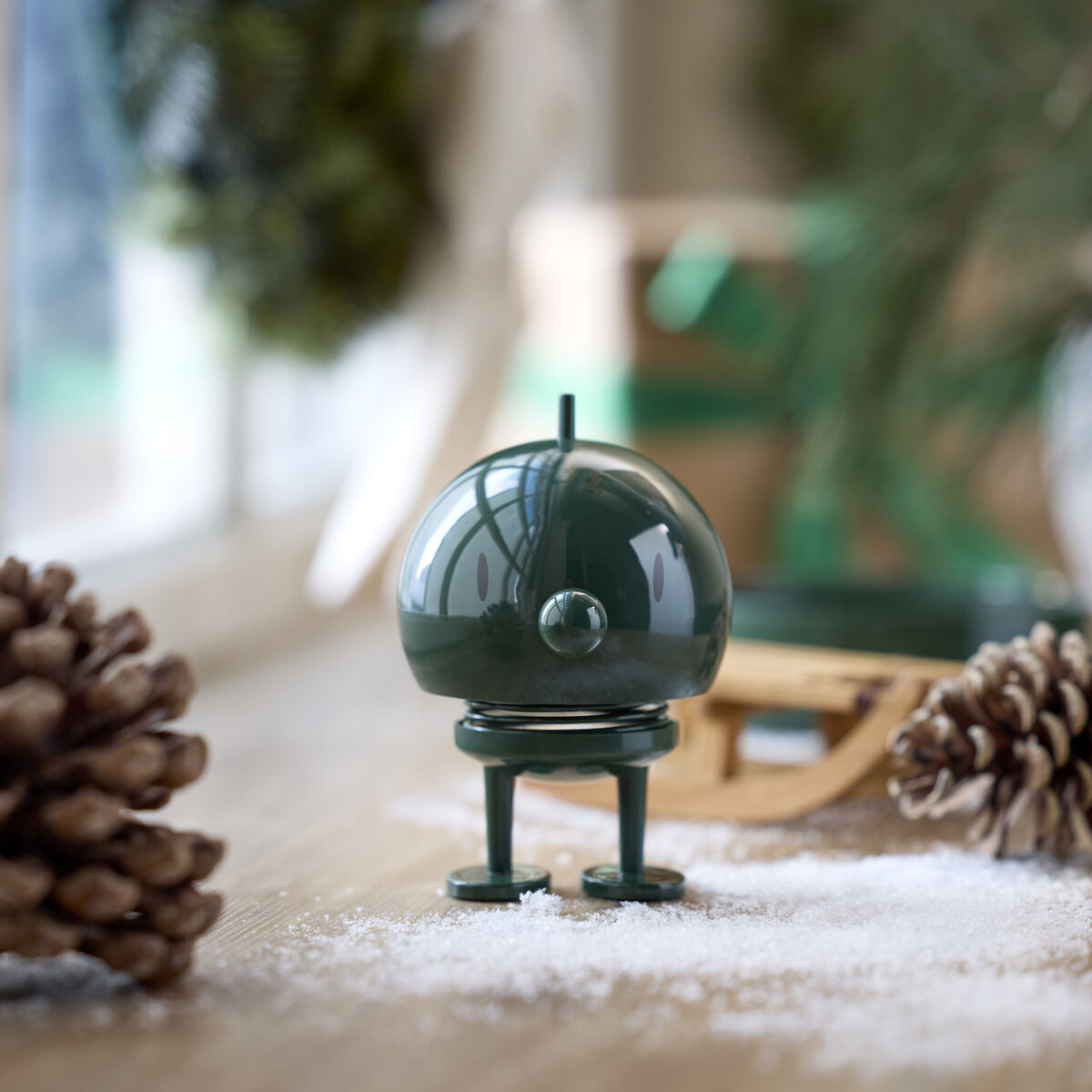 Ein Hoptimist Bumble Pine Green steht als dunkelgrüne, runde Designfigur mit federnden Beinen auf einem Holzboden in winterlicher Dekoration. Umgeben von Kiefernzapfen, Kunstschnee und einem Miniaturschlitten, nahe einem Fenster mit unscharfem Hintergrund.