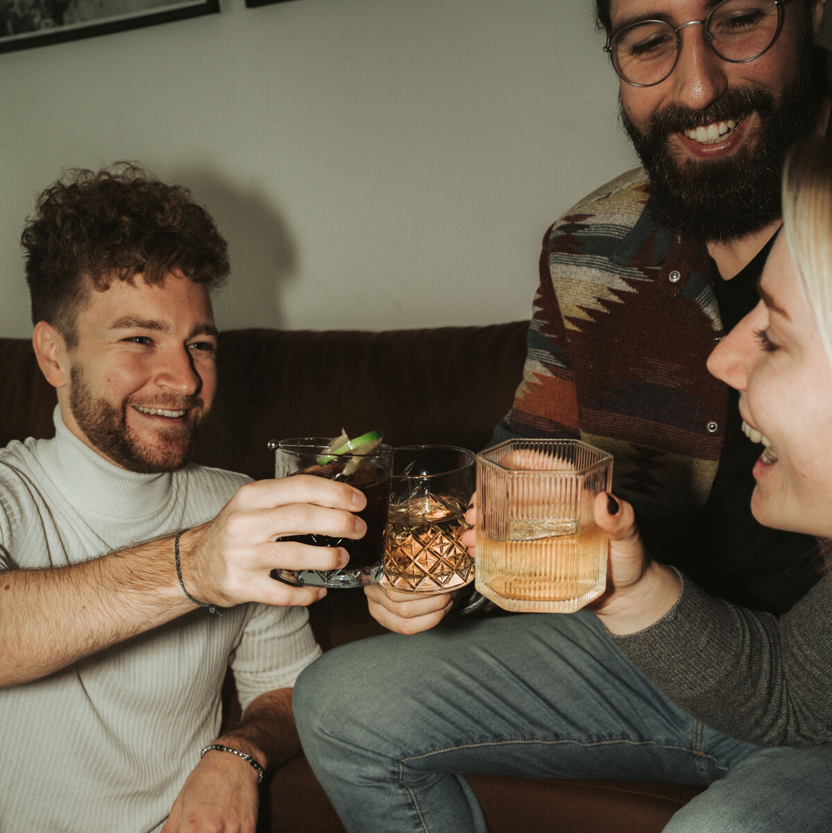 Drei erwachsene Freunde sitzen in einem gemütlichen Wohnzimmer und stoßen mit Gläsern an, die mit Ron Piet Rum, 10 Jahre gereift, gefüllt sind. Ein Tisch mit Gläsern und Snacks ist ebenfalls zu sehen. Alle lachen und genießen den Moment.