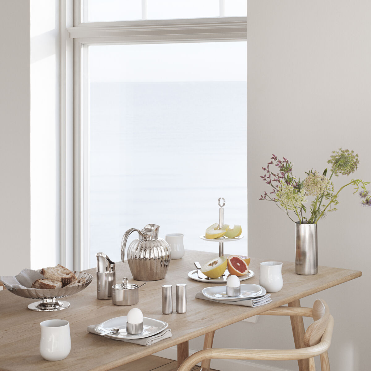 Bild eines einladend gedeckten Küchentisches mit einer weißen Zuckerdose mit Löffel aus der Kollektion 'Bernadotte'. Eingerichtet in einem hellen Raum mit Sicht auf das Meer, ergänzt durch Vasen mit Blumen und weißen Essgeschirr.
