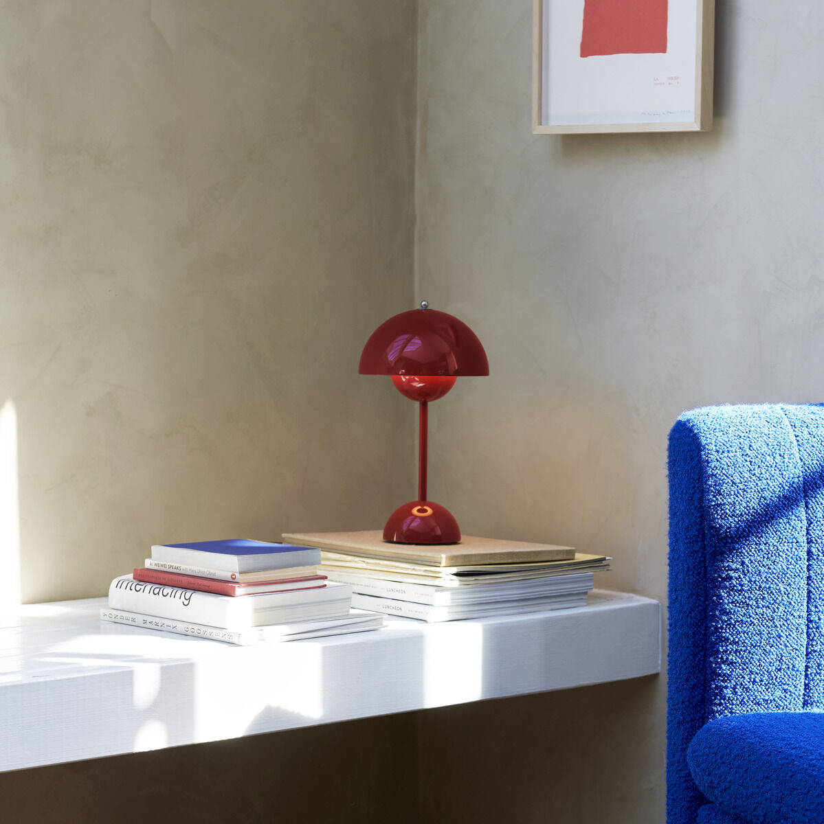 Bild einer modernen Inneneinrichtung mit einer roten Flowerpot VP9 Tischleuchte auf einem weißen Regal, daneben Bücher und ein Bild an der Wand. Ein blauer Sessel steht daneben auf einem hellen Holzboden.