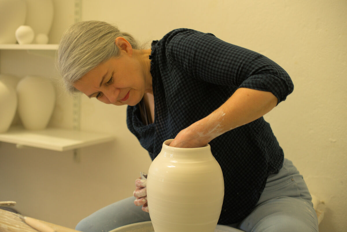 Eine Frau mit grauen Haaren in einem schwarzen Oberteil und hellblauen Jeans formt eine kleine, weiße Keramikvase mit ihren Händen. Im Hintergrund sind weiße Vasen auf Regalen zu sehen.