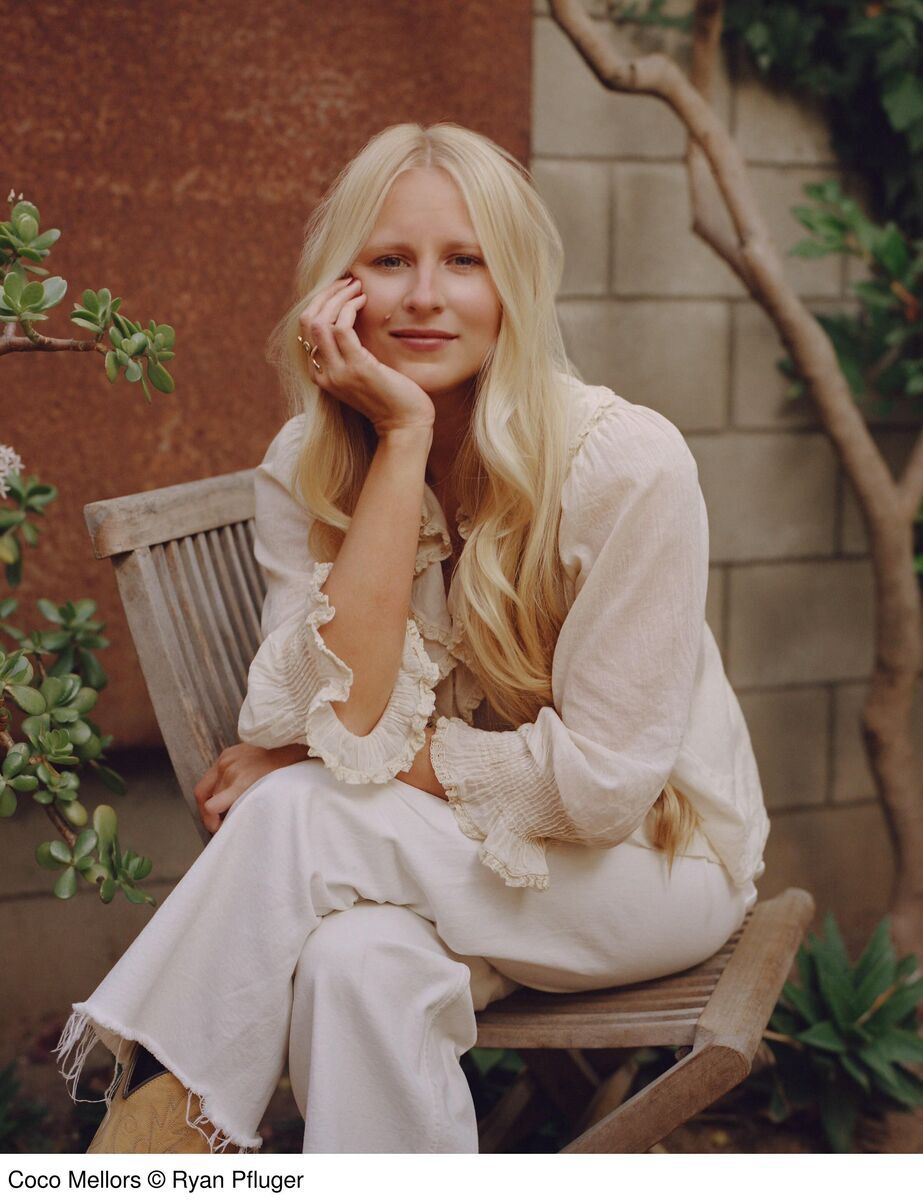 Eine Frau sitzt auf einem Holzstuhl und trägt weiße Kleidung. Im Hintergrund sind Pflanzen und eine Mauer zu sehen. Links unten ist der Text 'Coco Mellors © Ryan Pfluger' abgebildet.