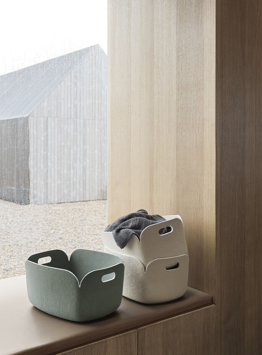 Drei minimalistische Aufbewahrungskörbe in verschiedenen Farben (grün, beige) aus Filzmaterial auf einer Fensterbank vor einem Holzfensterrahmen. Draußen ist ein modernes, mit Holz verkleidetes Gebäude bei regnerischem Wetter sichtbar.