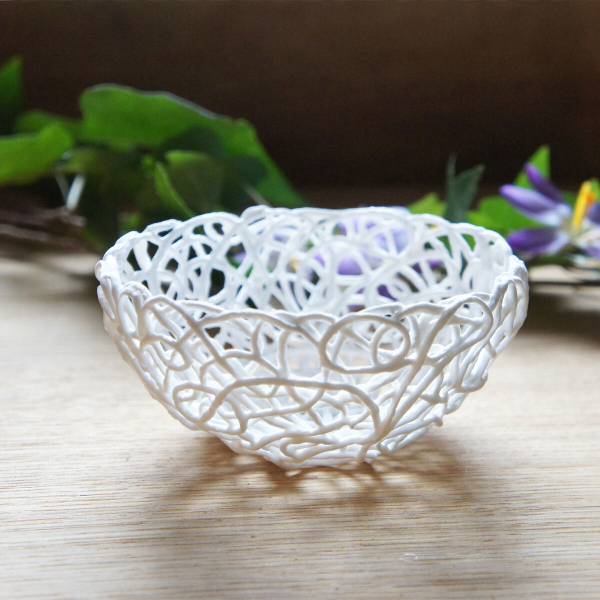 Eine weiße Porzellanschale mit filigranen Mustern steht auf einer hellbraunen Oberfläche. Im Hintergrund sind grüne und lila Blumenarrangements sichtbar.