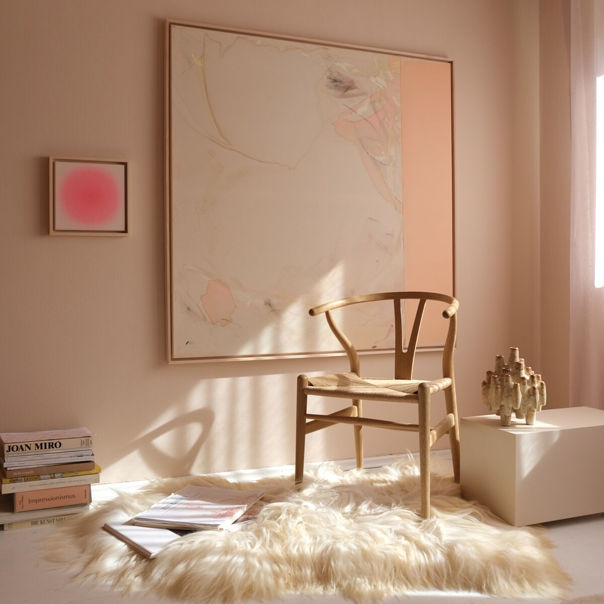 Eine moderne Einrichtung mit pastellfarbenen Wänden zeigt ein zeitgenössisches, quadratisches Wandbild in Rosa- und Beigetönen, einen naturfarbenen Stuhl aus Holz, ein flauschiges weißes Fell und helle Lichteinflüsse durch ein Fenster. Im Raum sind ein abstrakter Kunstdruck, Designbücher, eine Skulptur und ein weiterer kleineres Kunstwerk sichtbar. Das Gesamtbild wirkt minimalistisch und elegant.