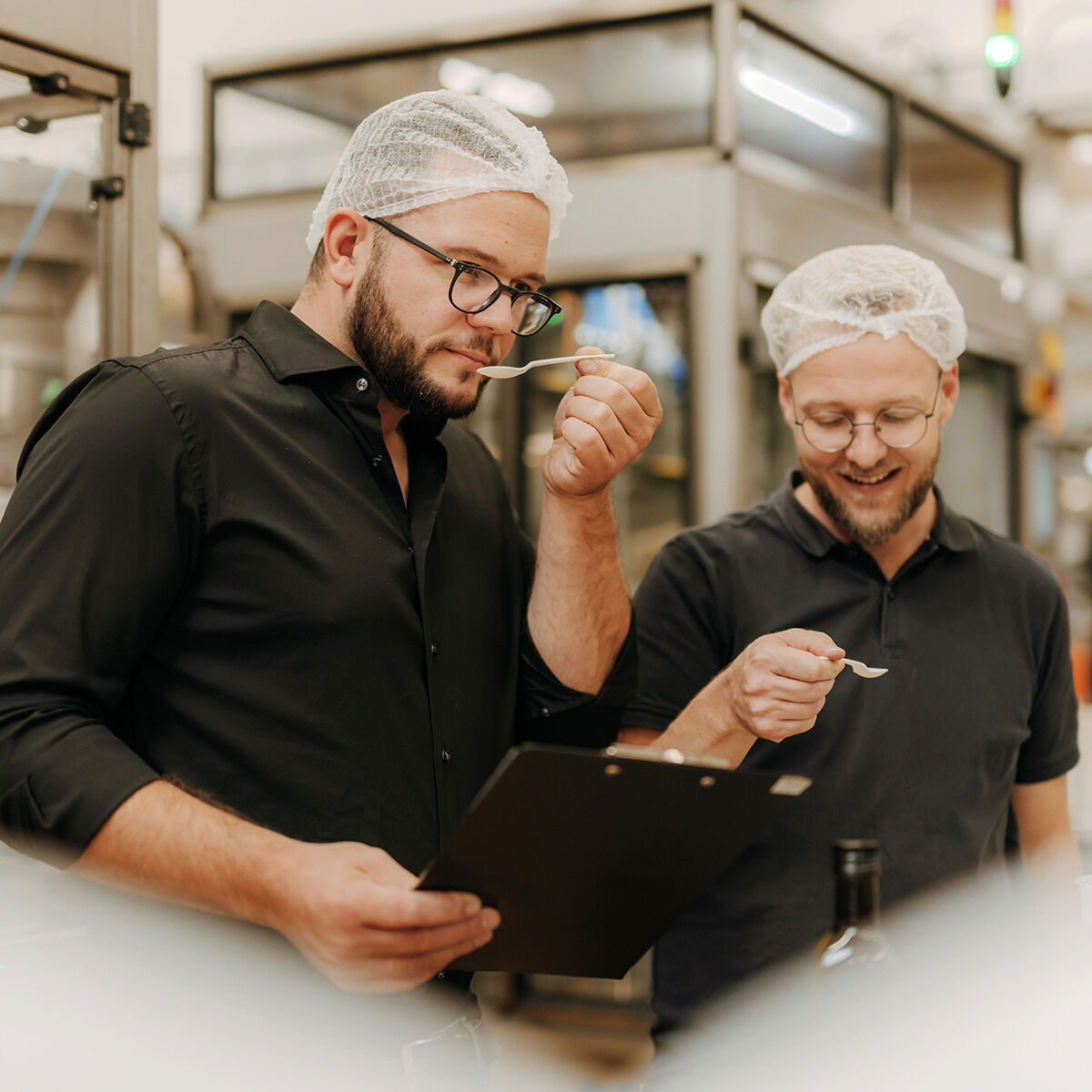 Zwei Männer mit Bart und Schutzhauben stehen in einer modernen Produktionshalle der Ölmühle Solling. Sie führen eine sensorische Qualitätskontrolle durch und probieren mit kleinen Löffeln Essigprodukte aus der ZEIT Präsentbox Jubiläumsessige. Einer hält ein Klemmbrett.