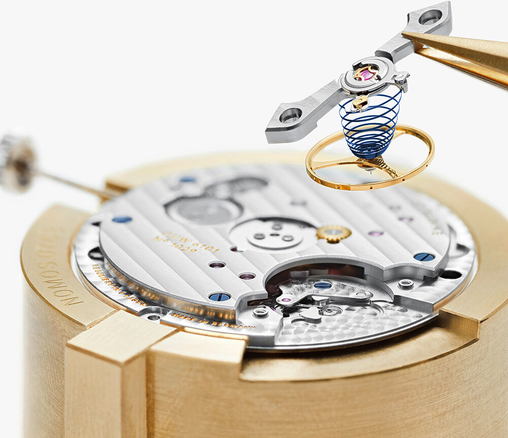 Detailaufnahme des exklusiven NOMOS Glashütte Uhrwerks der »Tangente Gold neomatik 38 Update DIE ZEIT«, montiert auf goldfarbener Halterung. Deutlich sichtbare gebläute Unruhspirale, goldene Brücken und rhodinierte Oberflächen spiegeln hochwertige Uhrmacherkunst wider.