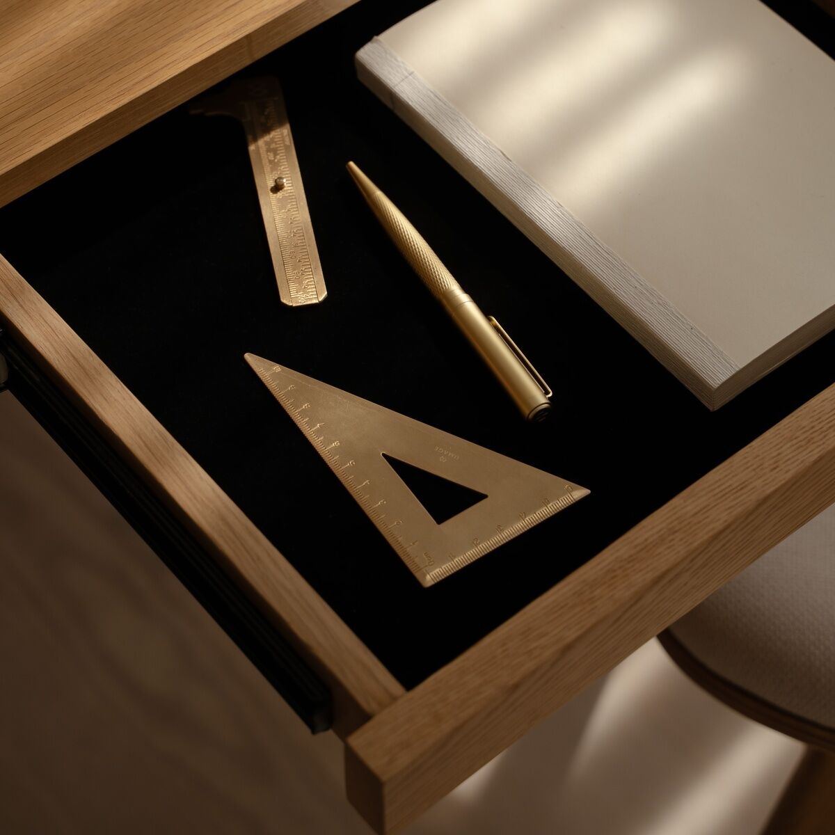 Die geöffnete Schublade des Umage Schreibtischs »Heart'n'Soul« mit schwarzer Innenfläche zeigt ein weißes Notizbuch, einen goldenen Stift, ein goldenes Dreieck und ein goldenes Lineal. Natürliches Licht fällt auf die Holzoberfläche. Design wirkt modern und elegant.