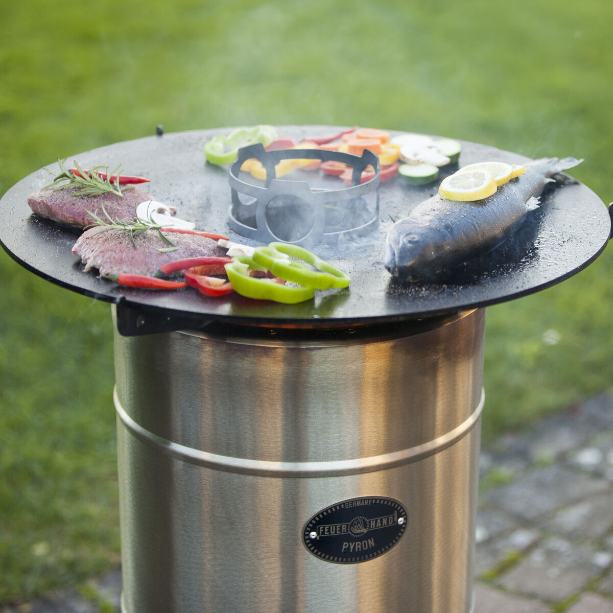 Eine Outdoor-Grillplatte von Pyron wird verwendet, um mehrere Lebensmittel gleichzeitig zu grillen, darunter Fisch, Fleisch und Gemüse. Die Grillplatte ist rund und steht auf einem silberfarbenen, zylinderförmigen Sockel. Im Hintergrund befindet sich Grünfläche.