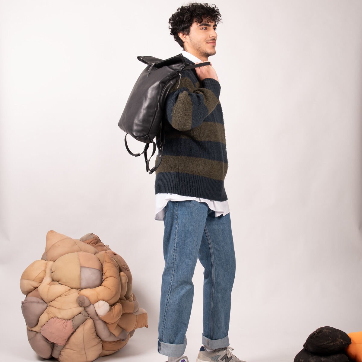Ein Mann in Jeans und einem gestreiften Pullover posiert mit einem schwarzen Leder Rucksack mit dem Namen »Nabuku« auf dem Rücken. Im Hintergrund sind künstlerische Dekoelemente sichtbar. Das Bild ist vor einem neutralen Hintergrund aufgenommen.