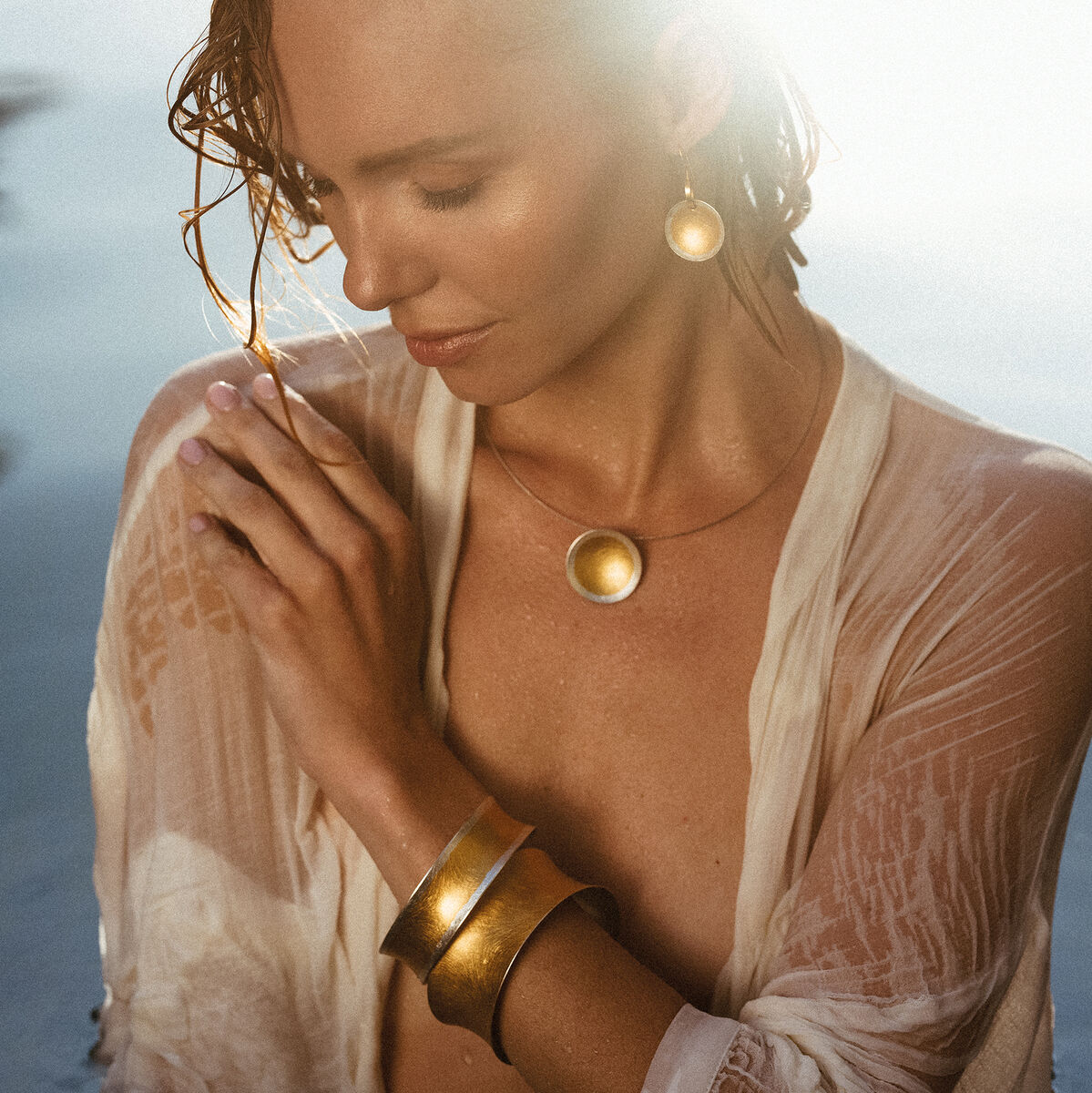 Eine Frau mit nassen Haaren trägt goldene Ohrringe »Sole«, eine Halskette und zwei Armbänder. Sie trägt eine halbtransparente weiße Kleidung und posiert nahe dem Wasser bei Sonnenuntergang.