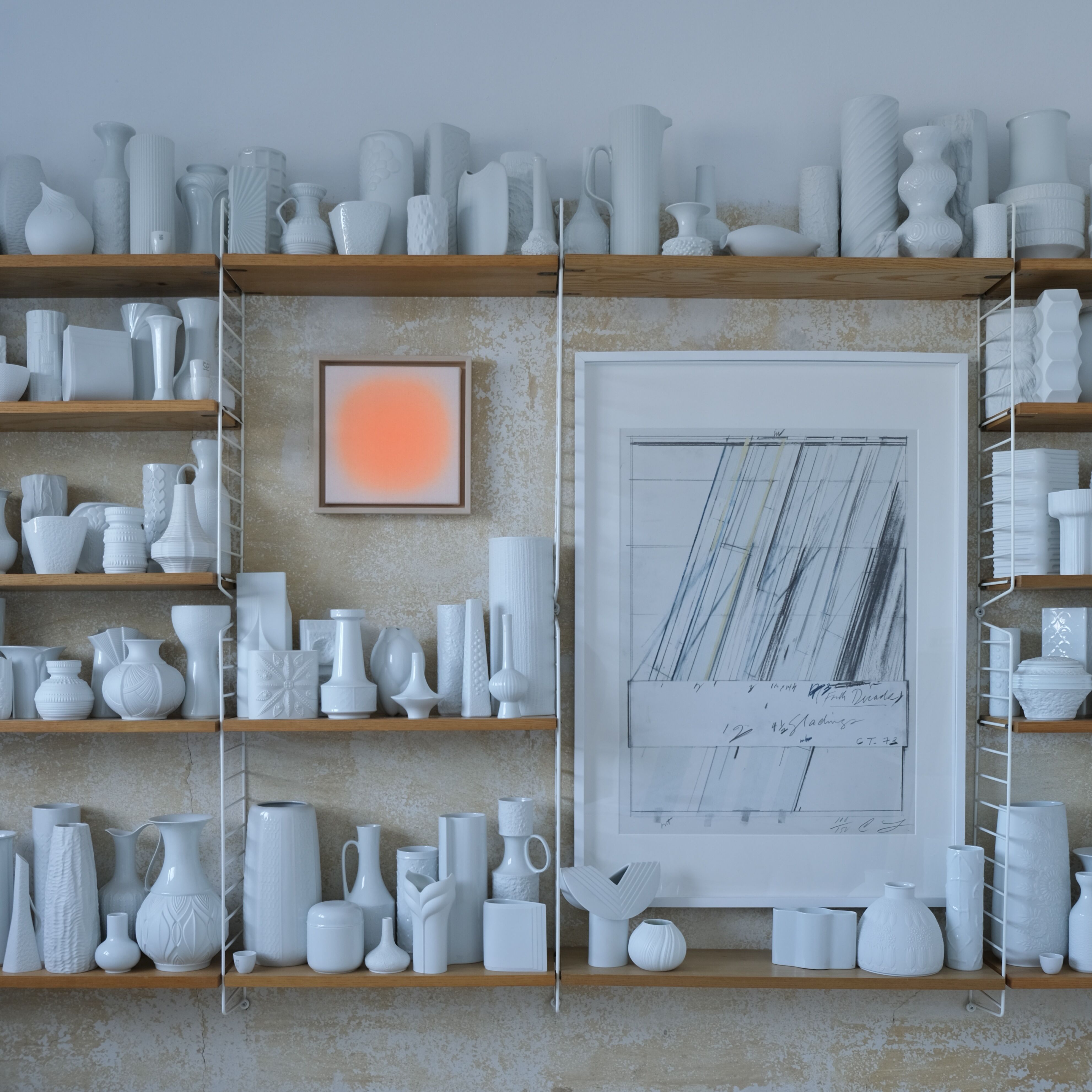 Moderne Wohnzimmerwand mit hellen Holzbrettern und weißen Keramikvasen sowie skulpturalen Objekten. Im Mittelpunkt hängt das Kunstwerk „ZEIT-Sonderedition »Sorbet«, Pink (30x30)“ von Anastasia Benko – ein quadratisches Bild mit sanft verlaufender pink-oranger Mitte. Daneben ist ein großes, grafisches Bild in Weiß und Blau zu sehen.