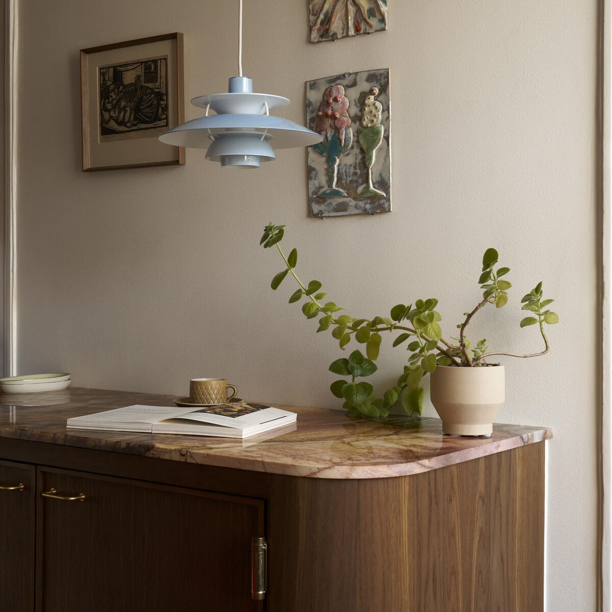 Im Zentrum hängt die Louis Poulsen Hängeleuchte »PH 5 320« in opal blau über einer Kommode mit Marmorplatte. An der Wand sind drei Kunstwerke, auf der Kommode steht eine Pflanze im Topf, eine Tasse und ein aufgeschlagenes Buch. Ruhige, zeitlose Atmosphäre.