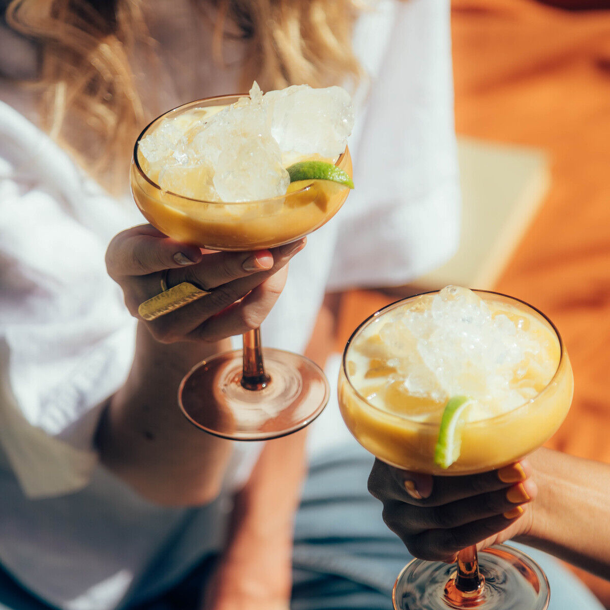 Zwei Personen stoßen im Freien mit Cocktails in eleganten Gläsern an, gefüllt mit gelbem Eierlikör, Eiswürfeln und Limettenscheiben. Im Hintergrund ist eine orangefarbene Decke und sommerliches Ambiente zu erkennen.