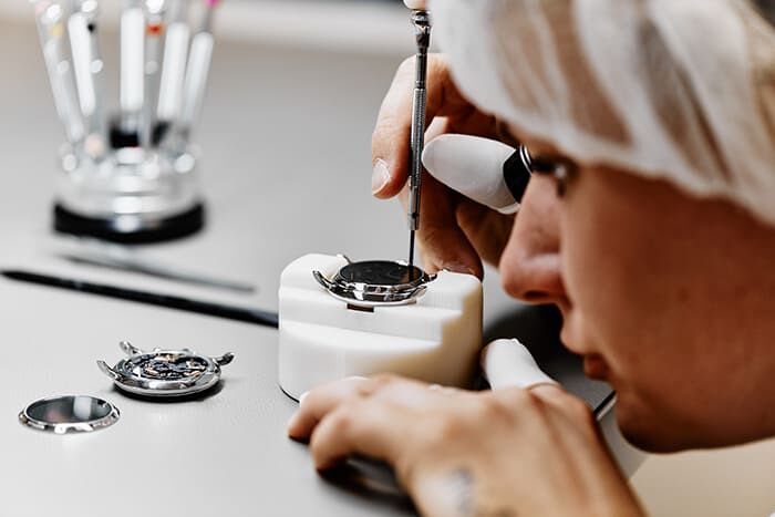 Ein gelernter Uhrmacher setzt ein Uhrwerk mit Präzisionswerkzeugen zusammen, umgeben von verschiedenen Uhrenteilen und Geräten.