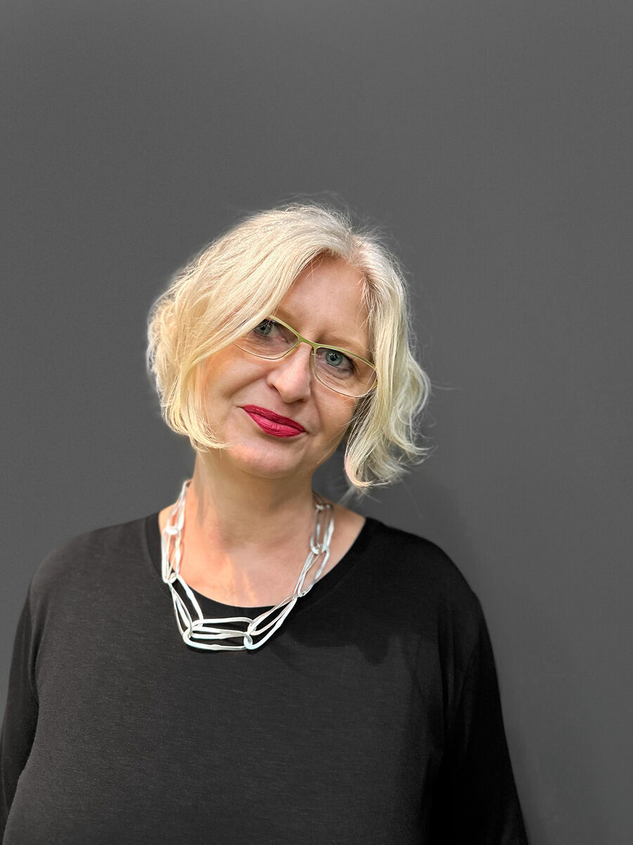 Eine elegante Frau mit blondem Haar trägt eine stilvolle Silberkette mit einem wellenförmigen Design. Der Hintergrund ist grau und die Frau trägt ein schwarzes Oberteil.