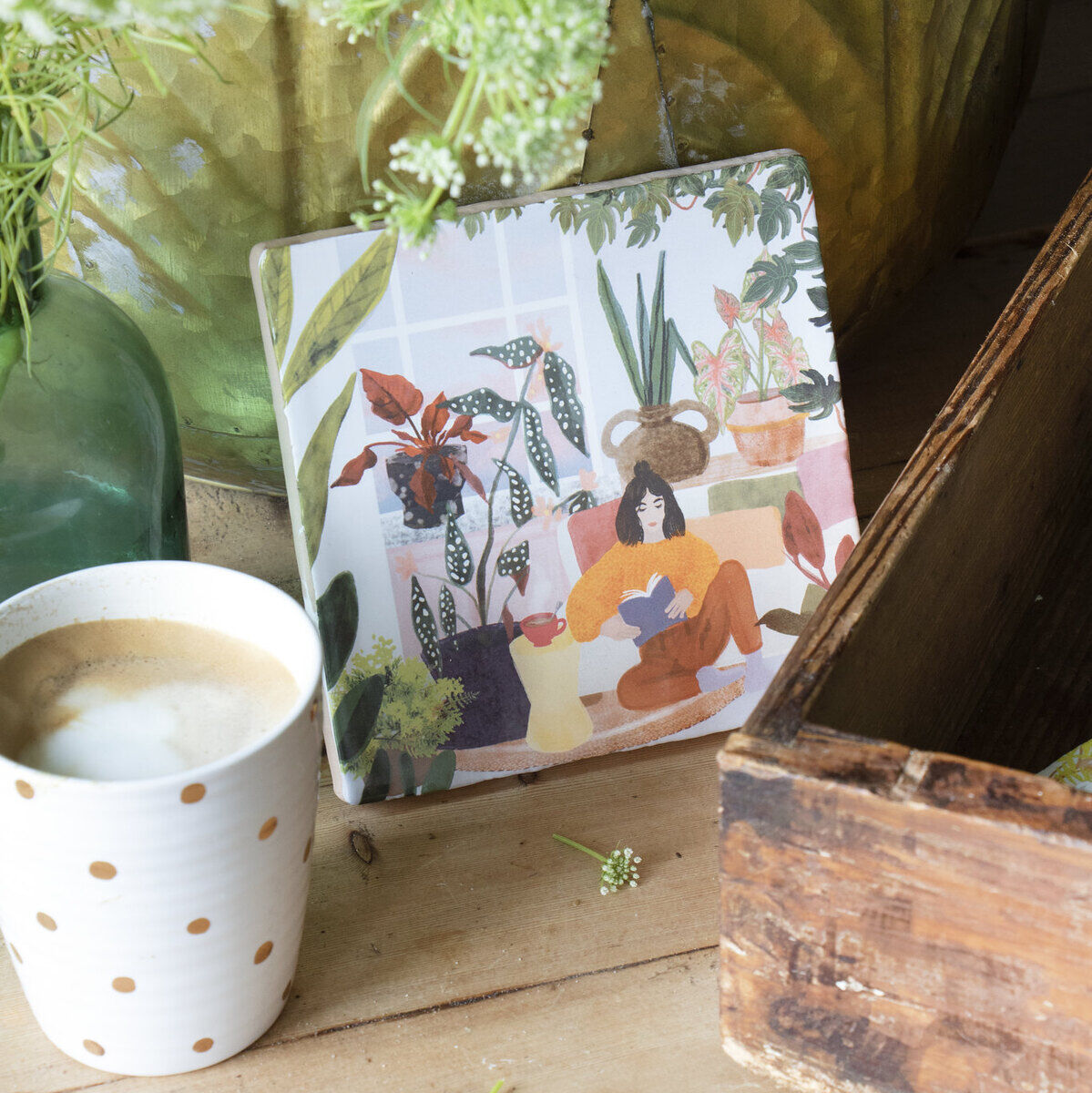 Eine gemütliche Szene mit Zimmerpflanzen, einem gerahmten Kunstwerk, das eine Frau mit Pflanzen darstellt, einem heißen Kaffee in einer gepunkteten Tasse und natürlichen Holzakzenten in einem hell beleuchteten Raum.