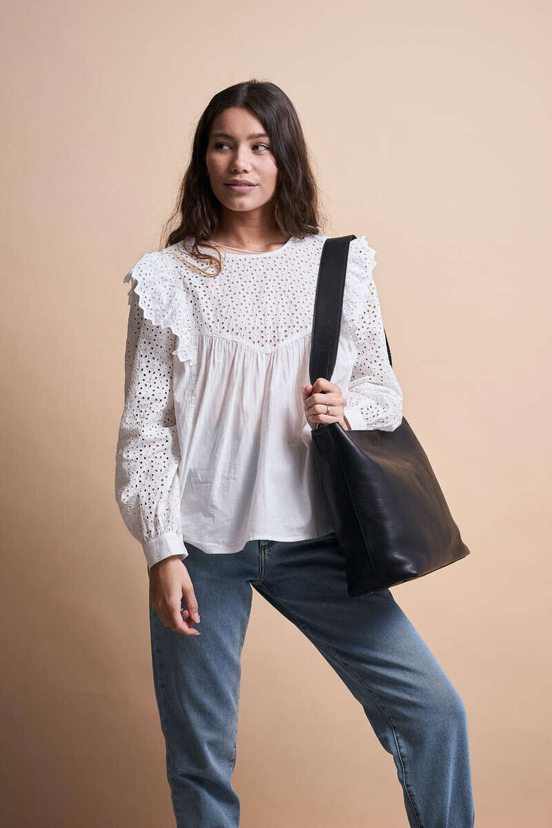 Eine junge Frau trägt eine schwarze Ledertasche über der Schulter. Sie trägt eine weiße, gehäkelte Bluse und eine blaue Jeans.