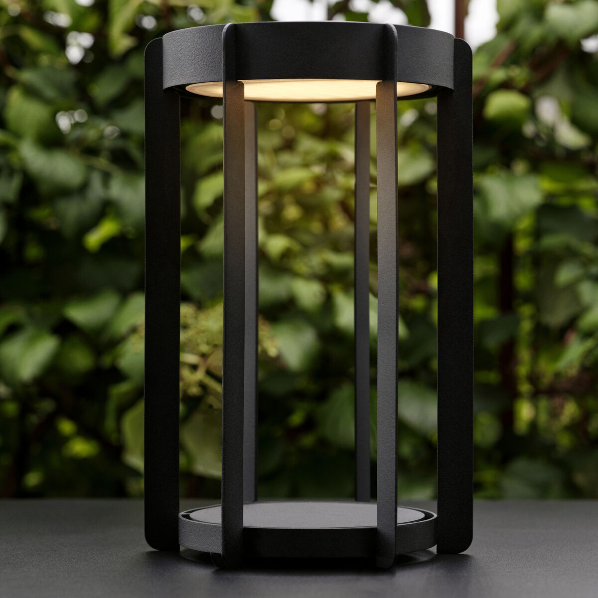 Eine schwarze LED-Laterne »Firefly«, abgebildet vor einer grünen Gartenkulisse. Die Laterne hat ein modernes, schlichtes Design mit geraden Linien und leuchtet warmes Licht ab.