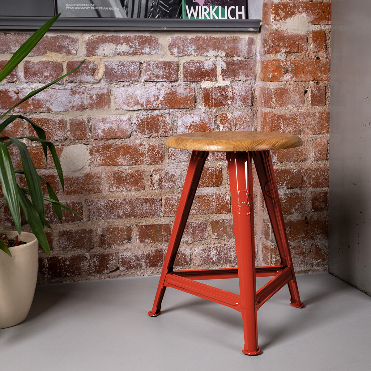 Ein Hocker der ZEIT-Sonderedition Rowac-Schemel »Red Brick« mit einem roten Metallgestell und einem hölzernen Sitz vor einer Ziegelwand, begleitet von einer Pflanze in einem Topf.