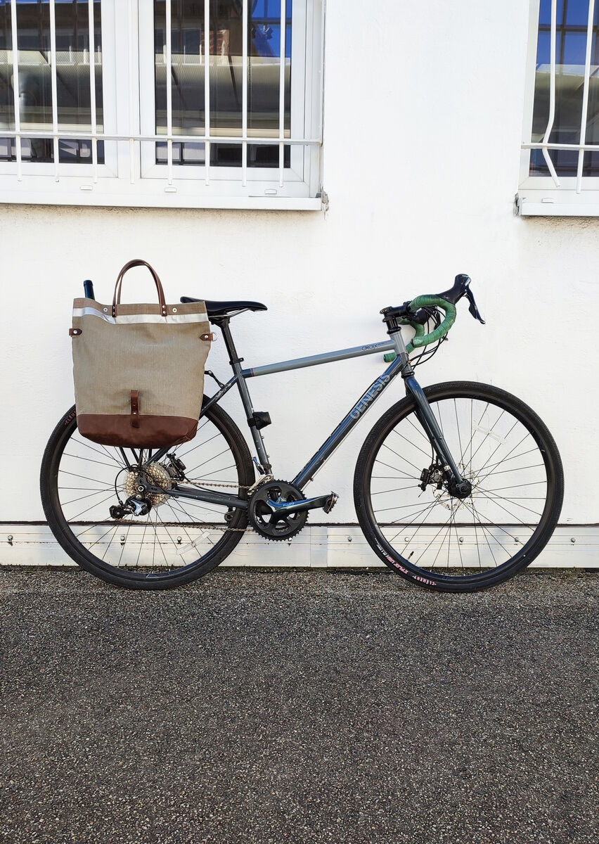 Ein graues Fahrrad ist an einer weißen Wand geparkt. An seinem Rahmen ist eine beige und braune Fahrradtasche befestigt, die wie eine Umhängetasche aussieht. Das Fahrrad hat eine grüne Lenkstange und schwarze Reifen.