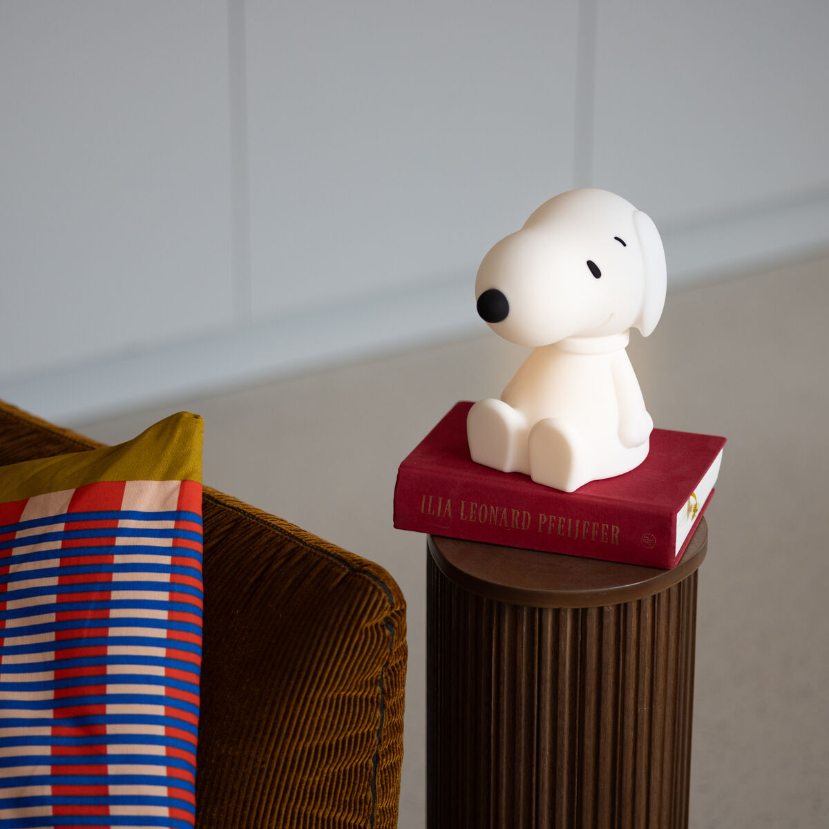 Das Bild zeigt ein Snoopy-Nachtlicht auf einem roten Buch, das auf einer geriffelten, braunen Säule steht. Neben dem Nachtlicht ist eine braune Couch mit einem Kissen, das mit bunten Streifen in Blau, Rot und Gelb gemustert ist, sichtbar. Der Hintergrund ist hell und minimalistisch gehalten.
