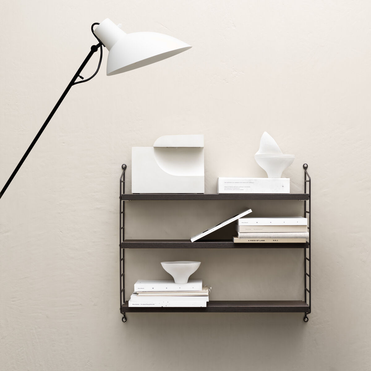 Ein modernes String Pocket Regal an einer beigen Wand. Das Regal ist aus schwarzem Metall und hält weiße Dekorationen und Bücher. Eine weiße Lampe ragt von der linken Seite ins Bild.