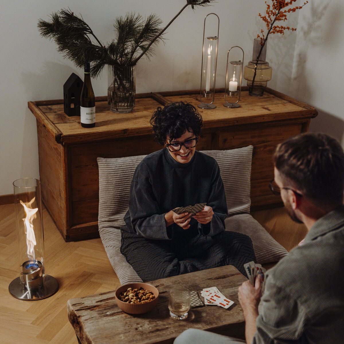 Ein Paar sitzt auf einer Couch und spielt Karten in einem gemütlich eingerichteten Wohnbereich mit einem Tischfeuergerät namens 'SPIN 1200' und mehreren dekorativen Kerzen und Pflanzen im Hintergrund.