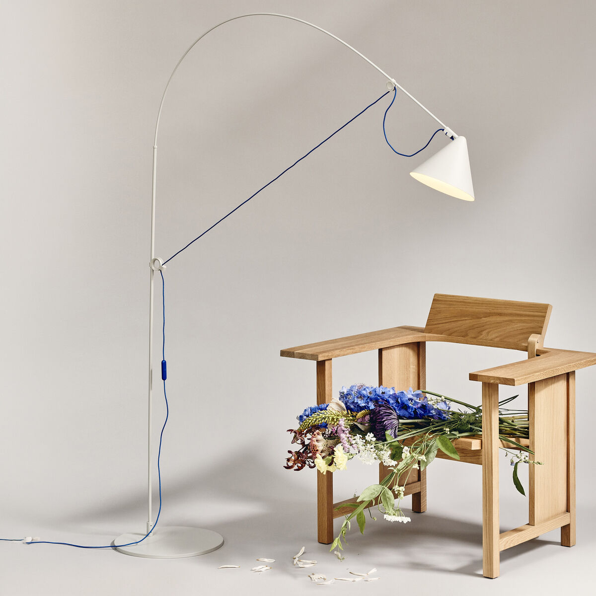 Stehlampe »AYNO« von Midgard in Silk-Grau und Ultramarin mit biegbaren Stab leuchtet stilvoll einen modernen Holzstuhl an, auf dem ein üppiges Blumenarrangement ruht. Die Szene ist minimalistisch und strahlt hochwertige, zeitgenössische Wohnästhetik aus.
