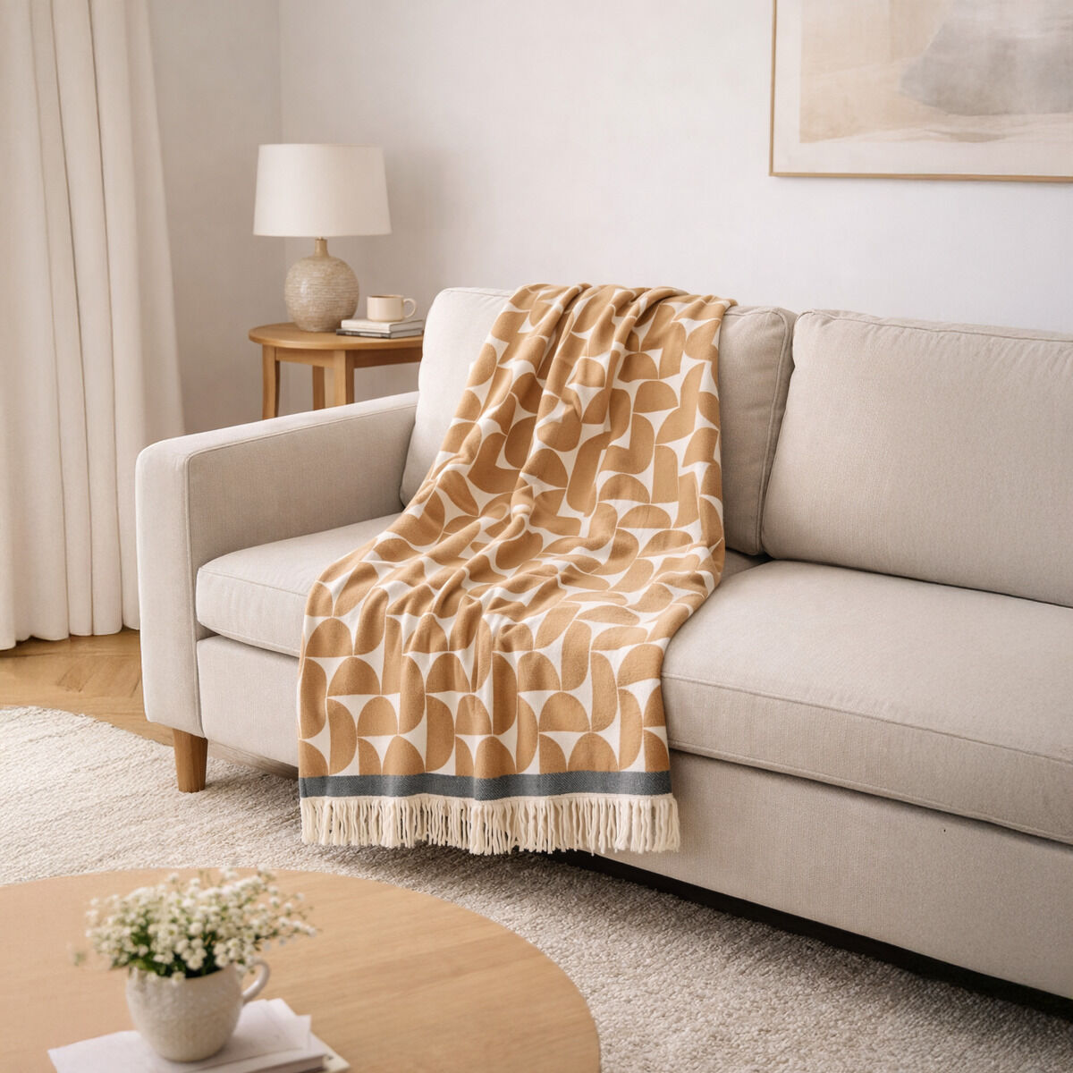 Auf einem hellgrauen Sofa liegt dekorativ die Baumwolldecke »Stockholm« von Eagle Products. Die Decke besticht durch ihr geometrisches Muster in Taupe und Creme, Fransen und farbige Bordüre. Daneben stehen ein Beistelltisch mit Lampe, Tasse und Deko in hell eingerichteter Umgebung.