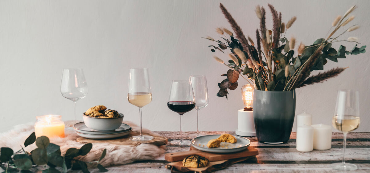 Ein stimmungsvoll gedeckter Tisch mit einer Auswahl an Weingläsern des Sommelier-Bundles »Vervino«, Kerzen, Snacks auf grauen Tellern, einem Strauß Trockenblumen in einer Vase und gemütlichem Kerzenlicht.