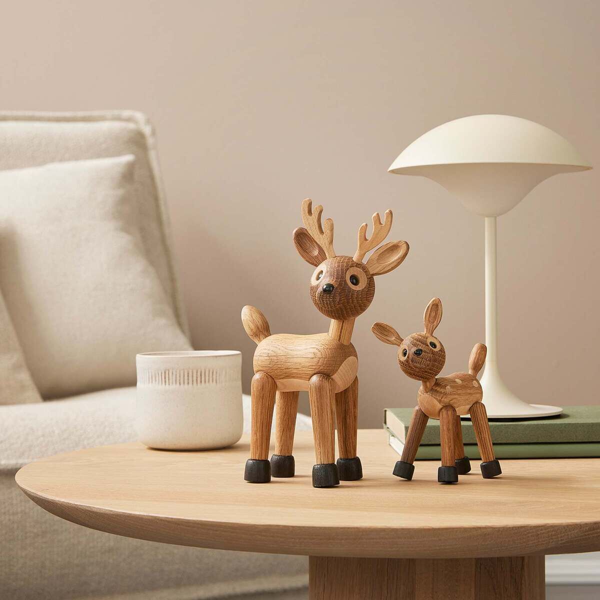 Zwei stilisierte Rentier-Holzfiguren, eine größere und eine kleinere, stehen dekorativ auf einem runden Holztisch in einem modernen, hell eingerichteten Wohnzimmer mit cremefarbenem Sofa, Lampe, Büchern und Kerze.