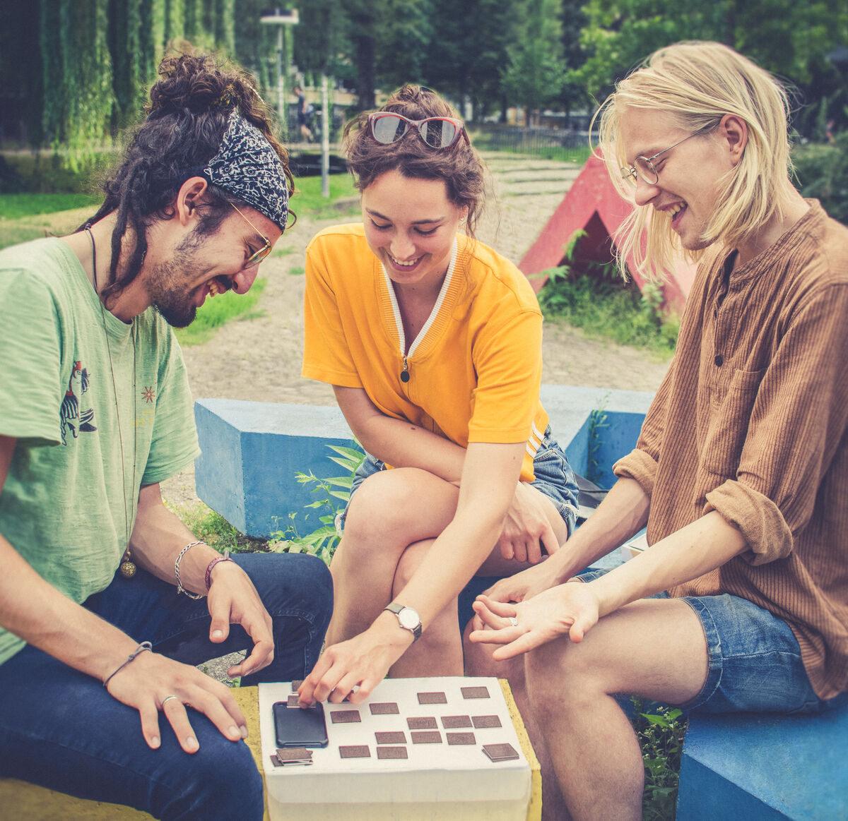 Drei junge Erwachsene spielen zusammen draußen ein Brettspiel mit Holzteilen aus der klang² Spielesammlung »Buchbinder Edition«. Sie sitzen auf bunten Bänken im Grünen und lachen.