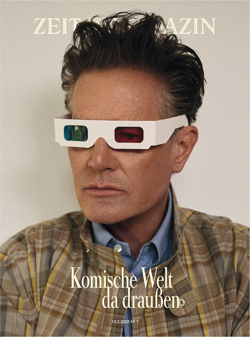 Cover der Zeitschrift DIE ZEIT 7/2025 mit einem Mann, der eine Retro-3D-Brille trägt. Der Hintergrund ist beige, während das Hemd in einem Karomuster aus Blau und Braun gestaltet ist. Der Schriftzug 'Komische Welt da draußen' ist in Weiß abgebildet.