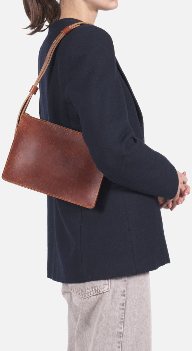 Eine Frau trägt eine schlichte Umhängetasche aus braunem Kernleder über der Schulter. Sie trägt einen dunkelblauen Blazer und helle Jeans. Die Ledertasche wirkt hochwertig, minimalistisch und edel, passend für Alltag oder Business.