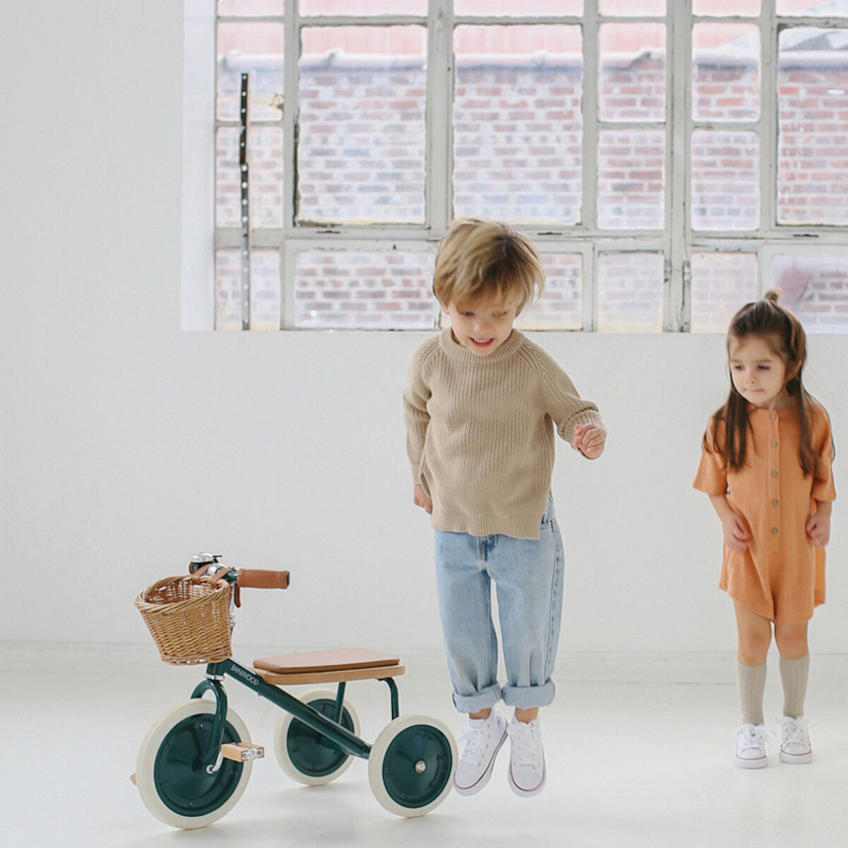 Zwei Kinder stehen in einem hellen Raum. Vor ihnen befindet sich ein Retro-Dreirad aus Holz und Metall mit einem kleinen Korb auf der Rückseite. Das Dreirad hat grüne Räder und braune Griffe. Im Hintergrund ist ein großes Fenster mit einem Blick auf eine Ziegelmauer zu sehen.