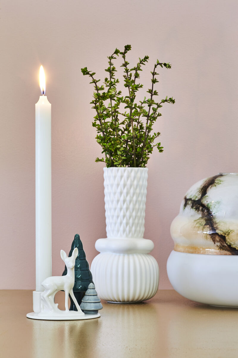 Kerzenständer mit brennender Kerze, Porzellanvasen und stehender Porzellanfigur von Bambi mit kleinen Tannenbäumen im Hintergrund vor einer pastellfarbenen Wand.