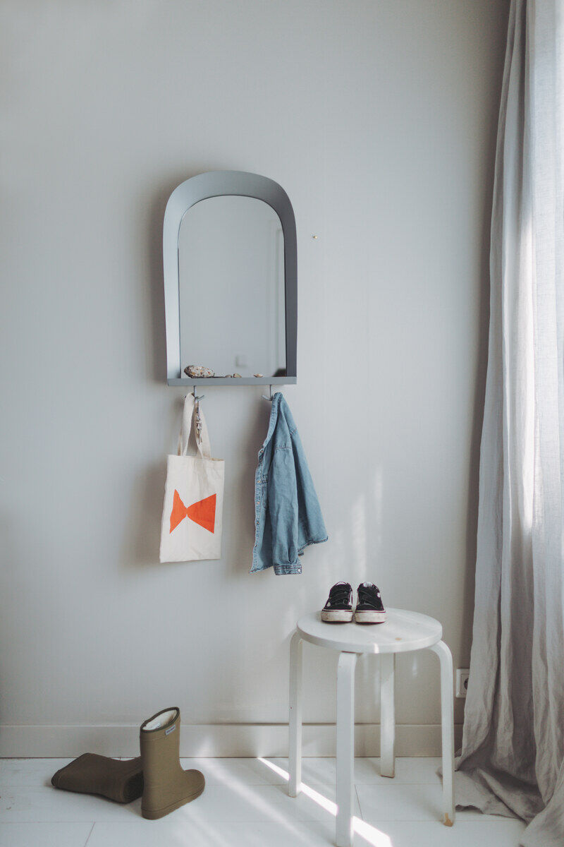 Ein modernes Kinderzimmer mit einem Wandspiegel, an dem eine Tasche und ein Kleidungsstück hängen. Darunter stehen ein grüner Gummistiefel, ein weißer Hocker und schwarze Kinderschuhe.
