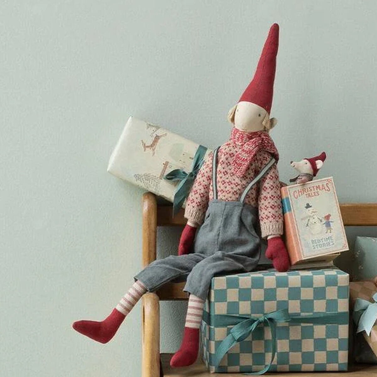 Auf einer Holzbank sitzen ein Maileg Kuscheltier »Weihnachtsmaus im Buch« mit roter Zipfelmütze und Strickpullover sowie mehrere pastellfarbene Geschenkpakete. Darunter und daneben stehen kleine, liebevoll gestaltete Miniaturmöbel und Figuren. Sanfte Farben und festliche Stimmung.