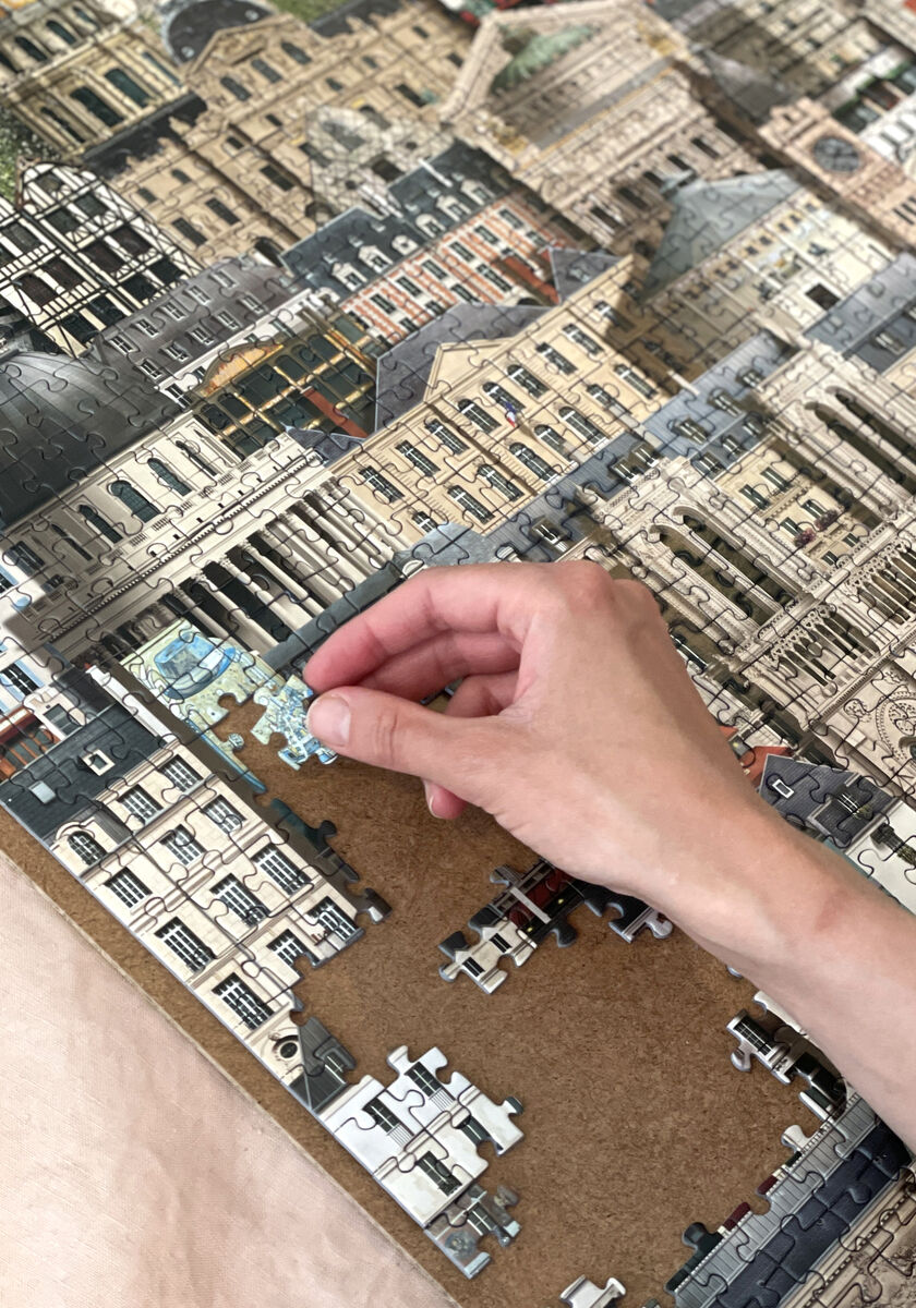 Eine Hand fügt ein Puzzlestück in ein fast fertiges 1000-teiliges Puzzle ein, das eine detaillierte Stadtansicht von Paris mit historischen Gebäuden zeigt.