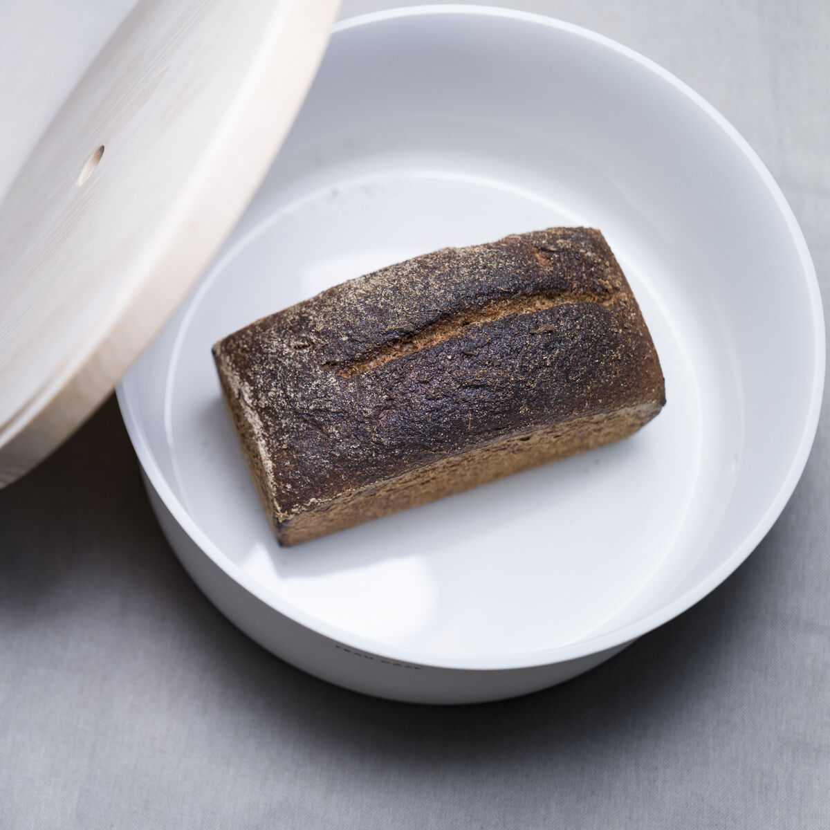 Detailreiche Aufnahme des Brottopfs „Brot & Brett“ von Frau Caze (Design: Carolin Zeyher) aus weiß mattem Porzellan mit massivem Eschenholzdeckel, geöffnet mit einem halben Brotlaib darin. Schlichte, minimalistische Gestaltung auf hellgrauem Untergrund.