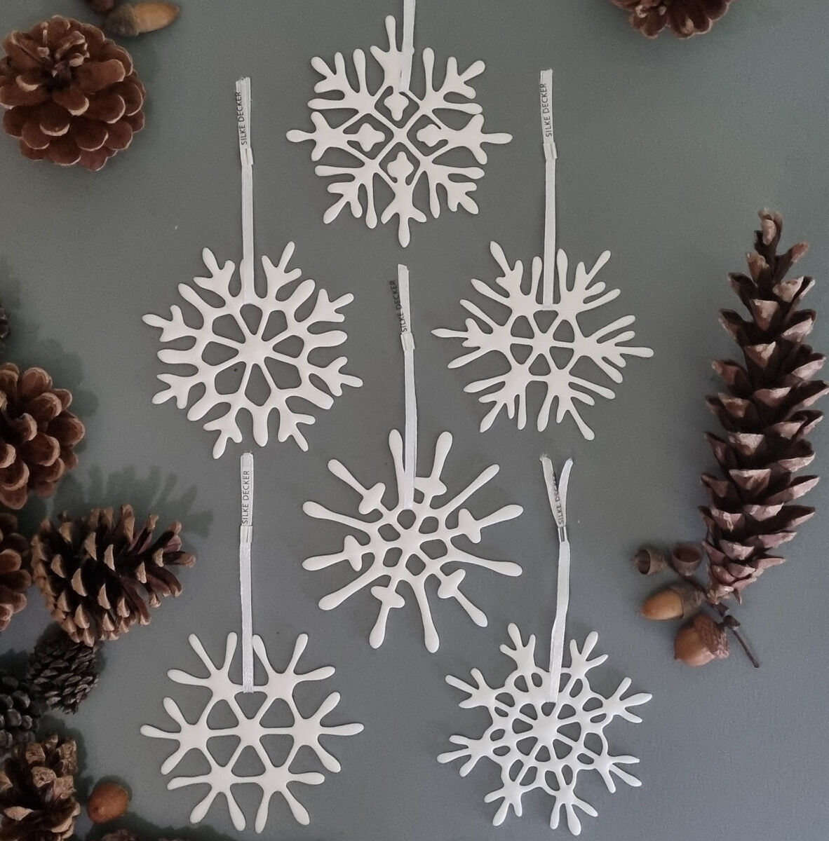 Sechs weiße, filigrane Schneeflocken-Anhänger aus Holz arrangiert auf einer grauen Oberfläche, umgeben von braunen Tannenzapfen und Eicheln. Die Anhänger sind als dekorative Winter- oder Weihnachtsdekoration geeignet.